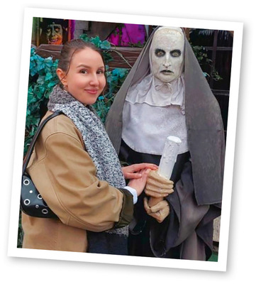 Eine Frau lächelt neben einer gruseligen Nonnenstatue, die eine Schriftrolle hält, mit Grünpflanzen und gruseligem Dekor im Hintergrund.