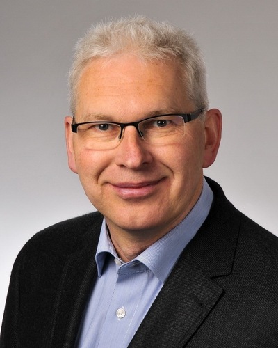 Ein Mann mittleren Alters mit kurzen grauen Haaren, Brille und Anzugjacke, der in die Kamera lächelt. - © Frank Eßmann