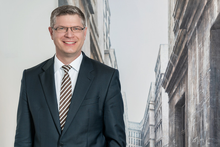 Ein lächelnder Mann in einem dunklen Anzug und einer gestreiften Krawatte steht vor einer Stadtlandschaft im Hintergrund. - © Leinemann Partner Rechtsanwälte