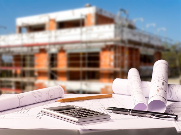 Baupläne mit Taschenrechner und einem Rohbau im Hintergrund