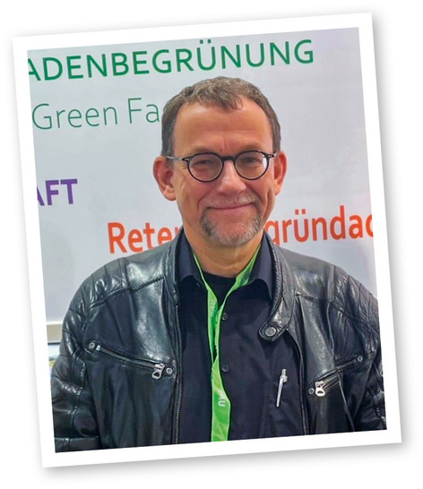 Ein Mann mit Brille und schwarzer Lederjacke lächelt vor einem Schild mit grün-orangefarbenem Text.