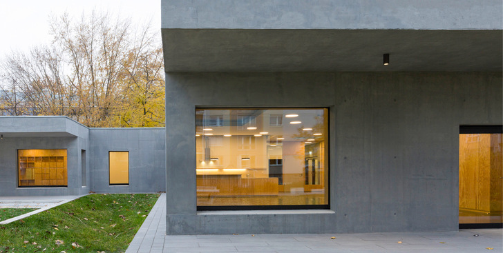 Modernes graues Betongebäude mit großen Fenstern und warmer Innenbeleuchtung, umgeben von Gras und Bäumen. - © Bild: Alexander Blumh﻿off