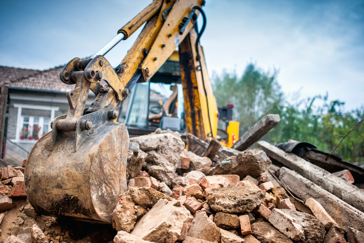 Ein gelber Bagger gräbt sich durch einen Haufen Schutt und zerbrochener Ziegel in der Nähe eines Hauses. - © Hoda Bogdan - stock.adobe.com