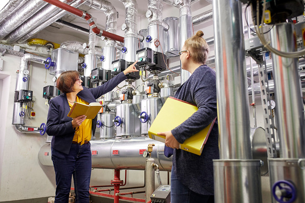 Zwei Personen mit Ordnern prüfen und besprechen Rohre, Ventile und Messgeräte in einem industriellen Maschinenraum. - © AMX Studio