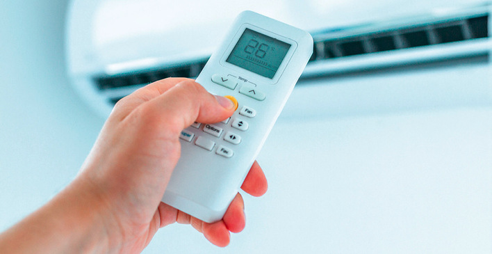 Hand hält eine auf 26 °C eingestellte Fernbedienung und zeigt auf eine an der Wand montierte Klimaanlage. - © Bild: Goffkein - stock.adobe.com