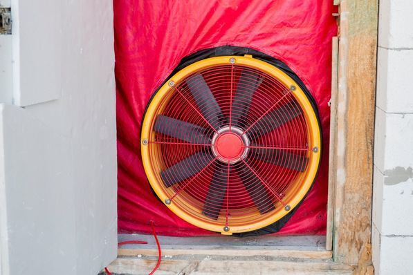 Großer Industrieventilator, mit rotem Stoff in einem Türrahmen befestigt, bläst Luft hinaus, umgeben von Baumaterialien. - © StockMediaProduction - stock.adobe.com