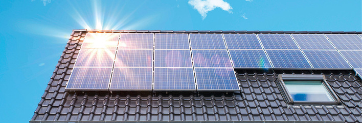 Solarmodule auf einem schwarzen Ziegeldach reflektieren das Sonnenlicht unter einem klaren blauen Himmel. - © Bild: Alessandro Terranova - stock.adobe.com