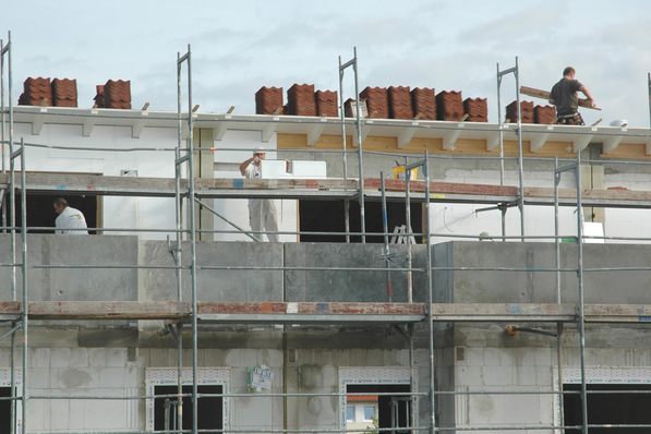 Drei Bauarbeiter arbeiten auf einem Gerüst an einem Gebäude, auf dem Dach sind Stapel von Dachziegeln zu sehen. - © VDPM
