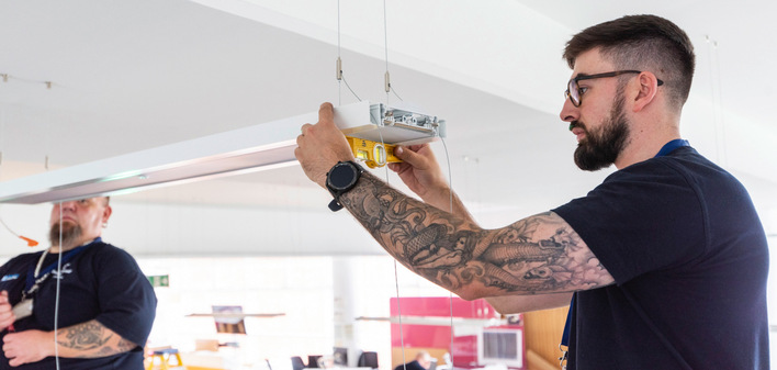 Zwei Techniker montieren in einer Industriehalle moderne LED-Leuchten, die Teil eines sensorgesteuerten und funkvernetzten Lichtsystems sind.